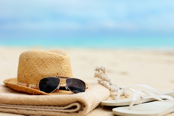 Sunglasses, hat, blanket, and sandals laying on the beach, representing time off and relaxation.