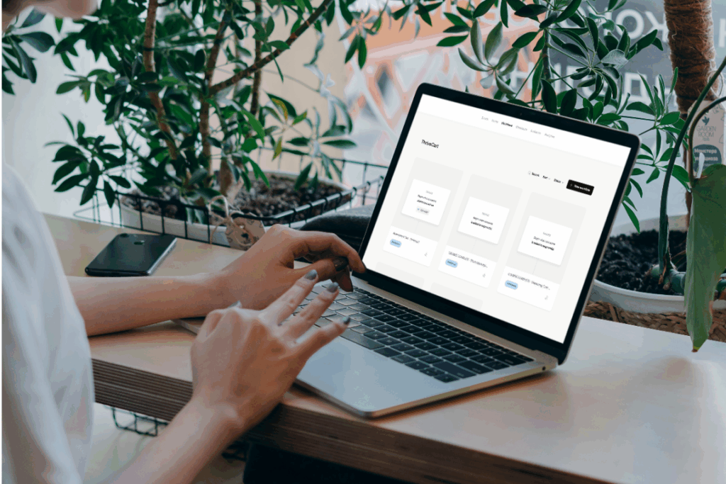 A person working on a laptop in a plant-filled workspace, with the Flodesk screen displaying Deb Mitzel Creative’s ThriveCart workflows, including a dashboard of digital product and automation tiles.