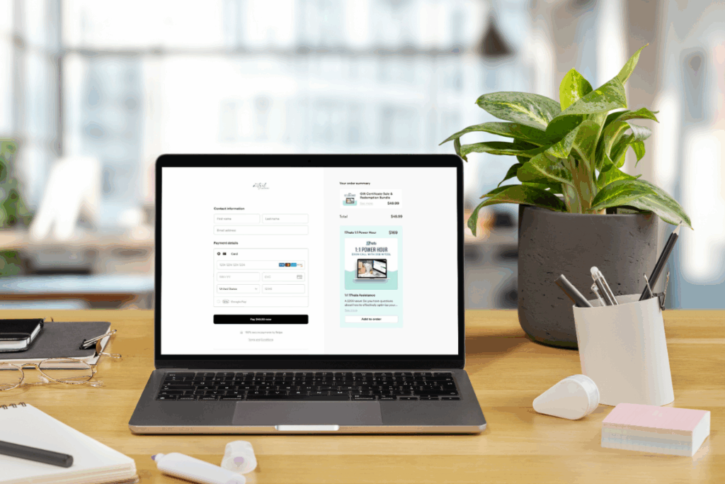 A laptop on a wooden desk displaying Deb Mitzel Creative's Flodesk checkout page for a digital product, including contact information fields, payment details, and an order summary with an optional add-on. The workspace includes a plant, notebooks, glasses, pens, and office supplies, with a bright blurred office in the background.